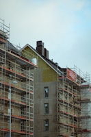 A building under construction is surrounded by scaffolding. The unfinished exterior walls have visible structural components and several windows, some covered with protective plastic. The overall scene has an industrial and developing feel, with a cloudy sky overhead.