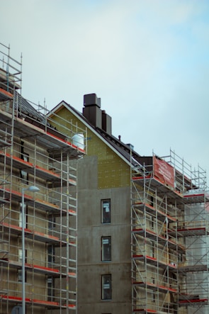A building under construction is surrounded by scaffolding. The unfinished exterior walls have visible structural components and several windows, some covered with protective plastic. The overall scene has an industrial and developing feel, with a cloudy sky overhead.
