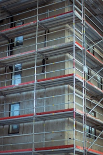 A multi-story building is under construction, surrounded by scaffolding. The structure is composed of metal railings and platforms that provide access to various levels. The building itself appears unfinished, with exposed concrete and visible windows.