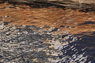 A chaotic interplay of reflected light creates an abstract pattern on the surface of water. The image captures the distortion of various colors, including brown, black, and white, in wavy motion.