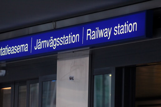 A sign displaying the word 'Railway station' in English, as well as in Finnish and Swedish (Rautatieasema and Järnvägsstation). The sign is mounted above likely a glass or metal structure, possibly an entrance.