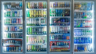 A well-lit row of refrigerated display cases filled with various bottled and canned beverages. The selection includes a wide range of drinks, organized neatly on shelves. Labels on the bottles and cans are visible, showcasing a variety of brands and flavors. The glass doors reflect some ambient light.