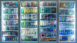 A well-lit row of refrigerated display cases filled with various bottled and canned beverages. The selection includes a wide range of drinks, organized neatly on shelves. Labels on the bottles and cans are visible, showcasing a variety of brands and flavors. The glass doors reflect some ambient light.