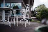 A patio scene with white metal chairs and tables on a wooden deck, surrounded by greenery and a modern building with glass windows in the background.