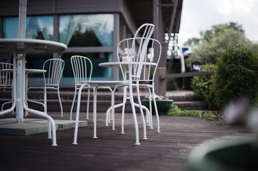 A patio scene with white metal chairs and tables on a wooden deck, surrounded by greenery and a modern building with glass windows in the background.