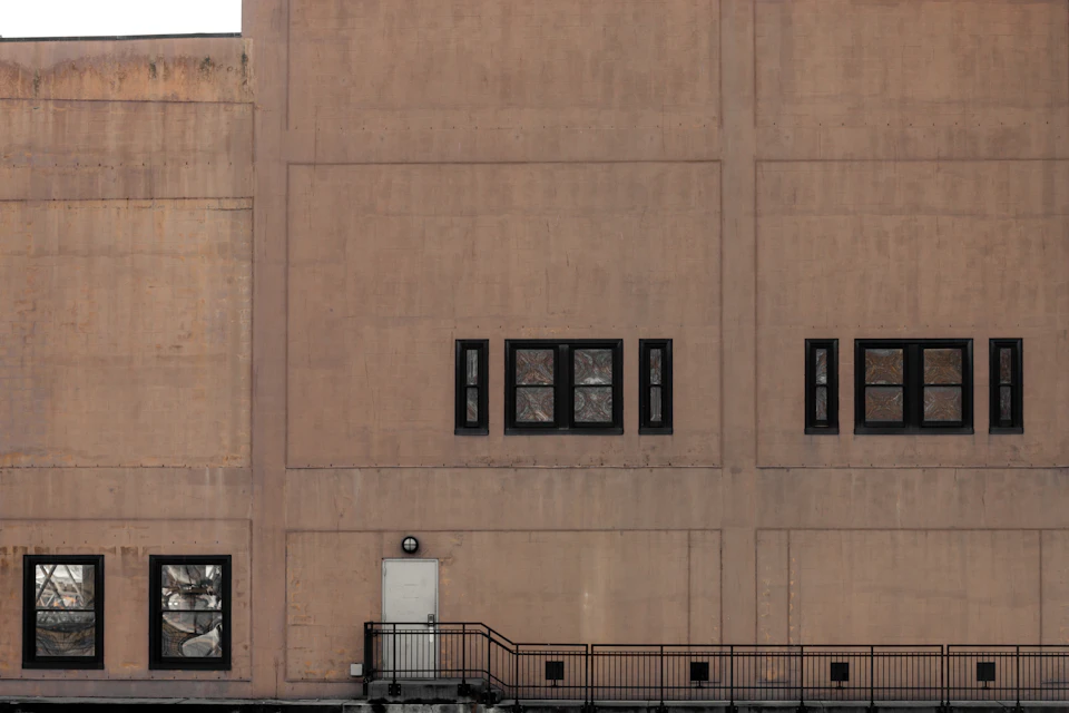 The image shows a large, light brown concrete building with multiple windows on its facade. The building has three rows of windows, each adorned with black shutters and white frames. A small door is located at the bottom right corner of the building. In front of the building, there is a metal railing that extends from the ground level to the top floor.