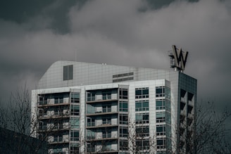 A modern high-rise building with a grid-like facade, featuring many windows and a prominent sign with the letter 'W' on the roof. The sky is overcast with dark, moody clouds, and bare tree branches are visible in the foreground.