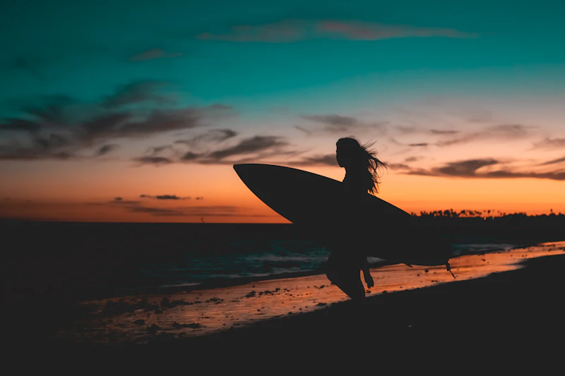 Silhouette of a surfer carrying her board along the beach at sunset