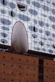 A modern architectural structure with a unique facade composed of irregular glass windows creating a wave-like pattern. The lower part is made of brick and features a large archway with small circular lights. People are visible on a terrace-like area between the brick and glass sections.