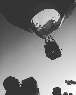 A black and white image featuring a hot air balloon from a low angle, showing the basket and the silhouette of a person inside. Several people on the ground look up at the balloon, some possibly capturing the moment with their phones. The sky is clear and contrasts with the dark silhouette of the objects.