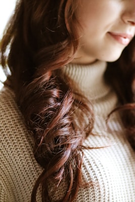 Close-up of a person's wavy hair and part of their face, focusing on luxurious, dark auburn curls. The individual is wearing a knitted, beige turtleneck sweater.