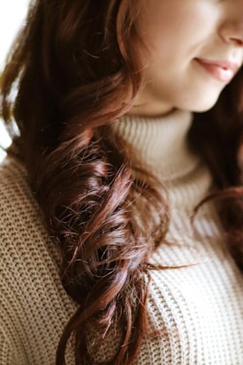Close-up of a person's wavy hair and part of their face, focusing on luxurious, dark auburn curls. The individual is wearing a knitted, beige turtleneck sweater.
