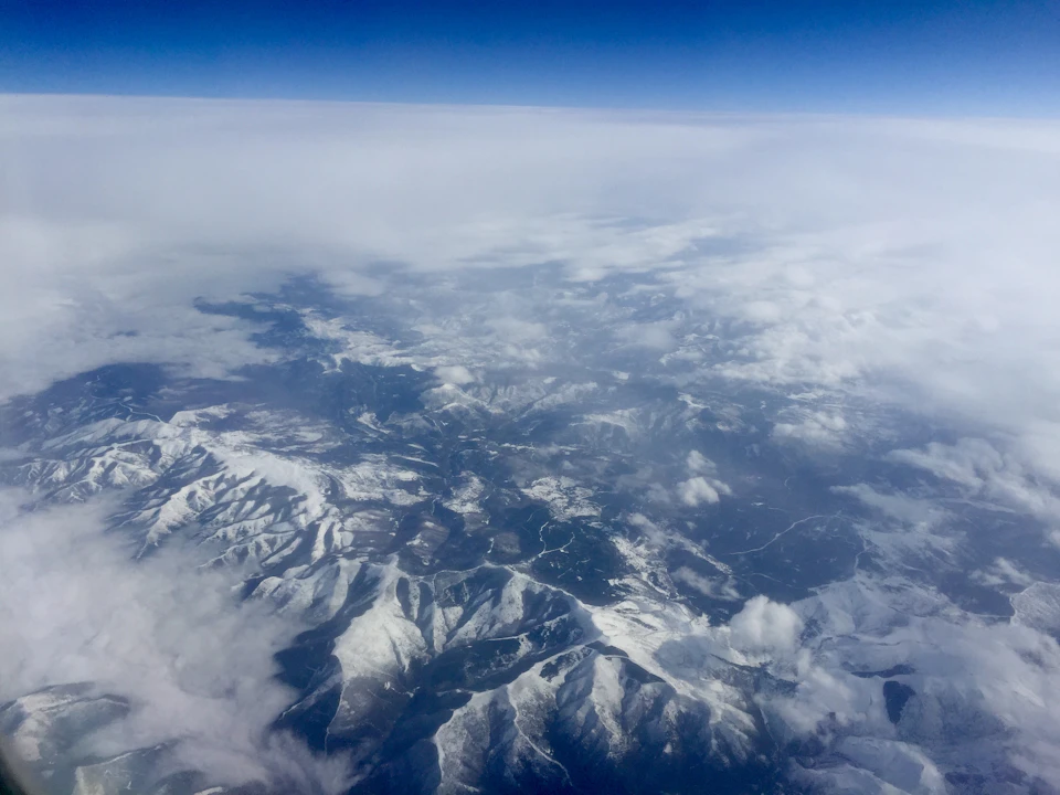 The image shows an aerial view of a mountain range with snow-capped peaks and valleys, set against a clear blue sky. The mountains are predominantly white due to their snow cover, while the valleys below have varying shades of green from vegetation. The perspective is from above, providing a comprehensive view of the landscape below.