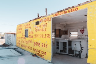 A dilapidated structure featuring bright yellow exterior walls covered with red graffiti, presenting survival instructions and warnings. The interior appears abandoned, with remnants of a kitchen visible and a heart symbol spray-painted inside.