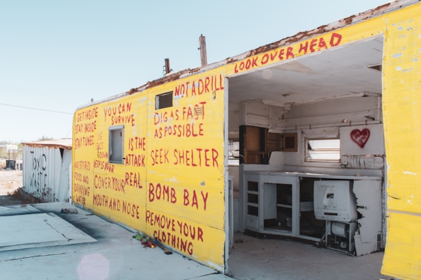 A dilapidated structure featuring bright yellow exterior walls covered with red graffiti, presenting survival instructions and warnings. The interior appears abandoned, with remnants of a kitchen visible and a heart symbol spray-painted inside.
