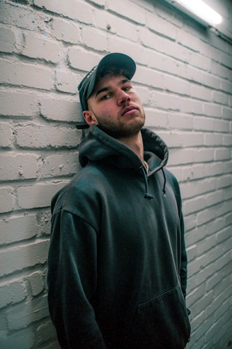 A person wearing a dark hoodie and a cap leans against a light-colored brick wall. The lighting is dim and casts soft shadows, creating a slightly moody atmosphere.