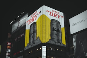 A large illuminated billboard featuring an advertisement for Asahi Super Dry beer dominates the scene with cans portrayed against a vibrant yellow background. The surrounding area includes other signs and advertisements, some partially visible, with a dark cityscape ambiance.
