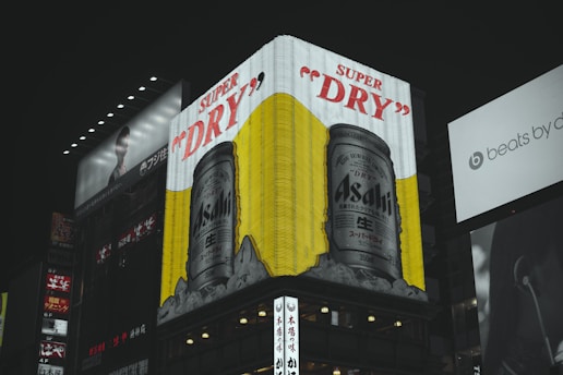 A large illuminated billboard featuring an advertisement for Asahi Super Dry beer dominates the scene with cans portrayed against a vibrant yellow background. The surrounding area includes other signs and advertisements, some partially visible, with a dark cityscape ambiance.