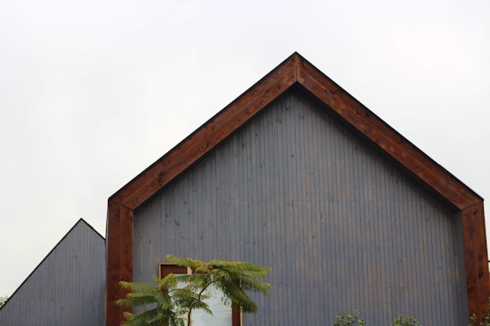A modern building with a distinct gable roof, featuring a wooden frame and vertical panels painted in muted gray. In the foreground, greenery including a tree with broad leaves is visible against a cloudy sky.