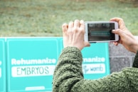 A person wearing a textured green sweater is holding a smartphone with both hands, capturing a photo of teal signs. The signs convey messages about rehumanizing embryos. The background appears to be an outdoor setting with grass.