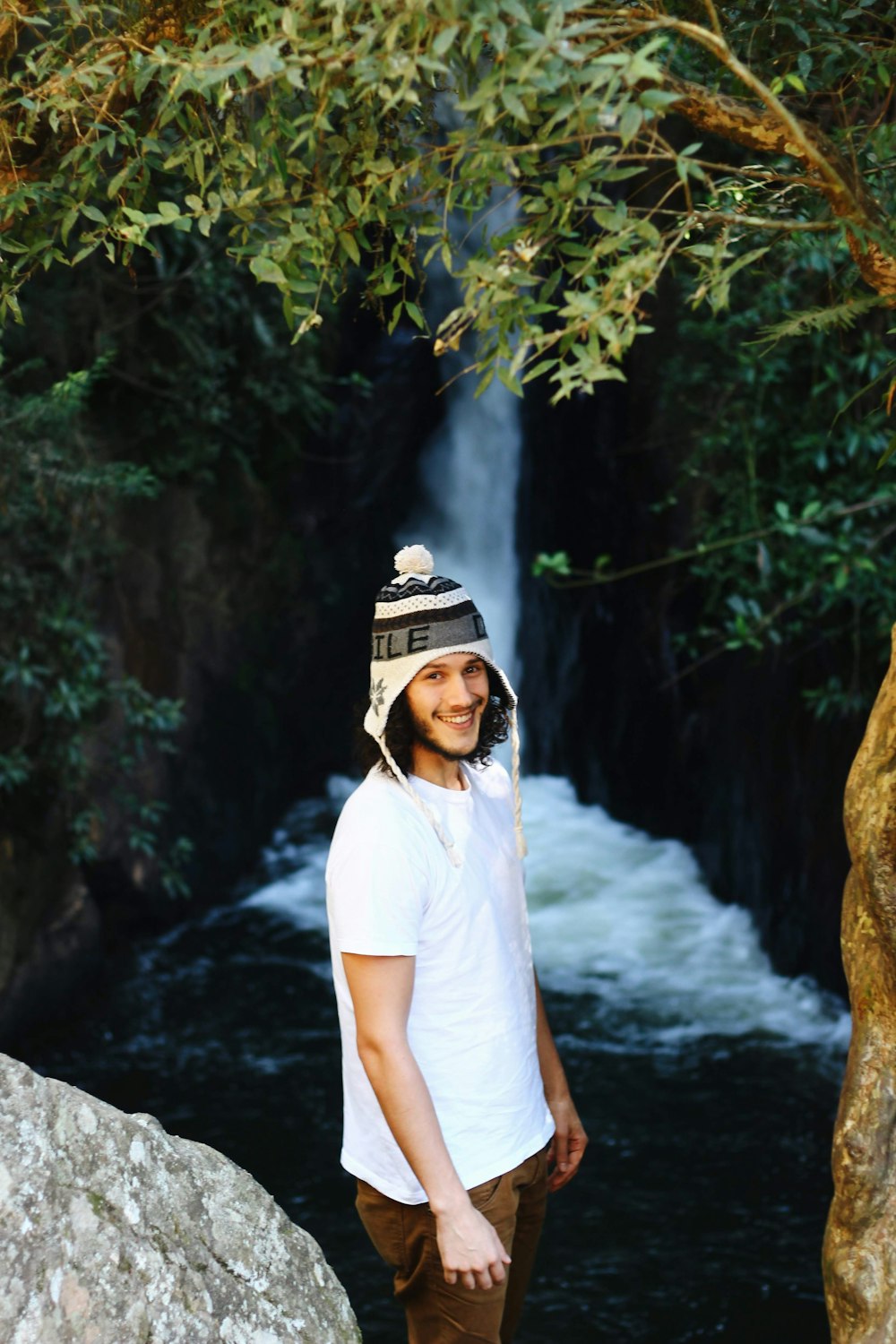 a man wearing a hat standing in front of a waterfall