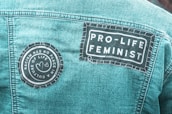 A denim jacket with two patches sewn onto the back. One patch reads 'Racism Has No Place in a Culture of Life,' featuring two heart shapes. Another patch says 'Pro-Life Feminist' in bold letters.