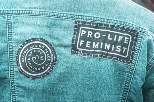 A denim jacket with two patches sewn onto the back. One patch reads 'Racism Has No Place in a Culture of Life,' featuring two heart shapes. Another patch says 'Pro-Life Feminist' in bold letters.