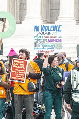 A group of people gathered in front of a building with large columns, holding signs advocating against violence and for women's empowerment. One sign reads 'NO VIOLENCE AGAINST' followed by different groups of people, written in colorful letters. Another sign urges to 'BREAK ALL the CHAINS!' and mentions the empowerment of women. The people are wearing casual clothing and some have orange shirts or jackets, and there is a uniformed officer present.