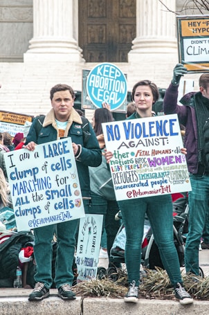 A group of people participating in a protest, holding signs advocating for pro-choice and other social justice causes. The scene is set on the steps of a governmental building, possibly a courthouse. The people are wearing warm clothing, suggesting it's a cooler day.