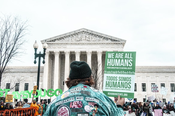 A group of people participate in a protest in front of a large, historic building with columns, possibly a government building. Many hold signs with various messages advocating human rights. A prominent sign in the foreground reads 'We Are All Humans' in both English and Spanish. The atmosphere suggests a peaceful demonstration.