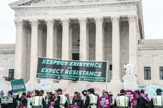 A group of people gather in front of a large, columned courthouse building holding various signs related to pro-life and pro-choice movements. The prominent sign reads 'Respect Existence or Expect Resistance.' Police officers are visible among the crowd.