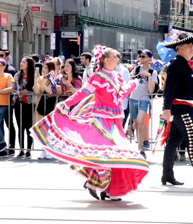 A woman dances joyfully in a vibrant, traditional dress featuring bright pinks and multicolored patterns. She is surrounded by a crowd of people holding flags and watching the performance. A man in a traditional outfit is nearby, wearing a decorated hat. The scene takes place outdoors in a lively setting.
