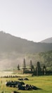 A serene landscape features a group of people gathered with tents and a vehicle on a green field. In the background, there are multi-tiered pagodas and lush mountainous terrain enveloped in a light mist, under a clear sky.