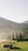A serene landscape features a group of people gathered with tents and a vehicle on a green field. In the background, there are multi-tiered pagodas and lush mountainous terrain enveloped in a light mist, under a clear sky.