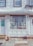 A storefront with a display window featuring an eclectic collection of humorous and quirky signs. The façade is simple with white siding and framed windows, showing some aging. The sidewalk is made of uneven stone tiles, reflecting a rustic charm.