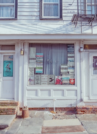 A storefront with a display window featuring an eclectic collection of humorous and quirky signs. The fa&ccedil;ade is simple with white siding and framed windows, showing some aging. The sidewalk is made of uneven stone tiles, reflecting a rustic charm.
