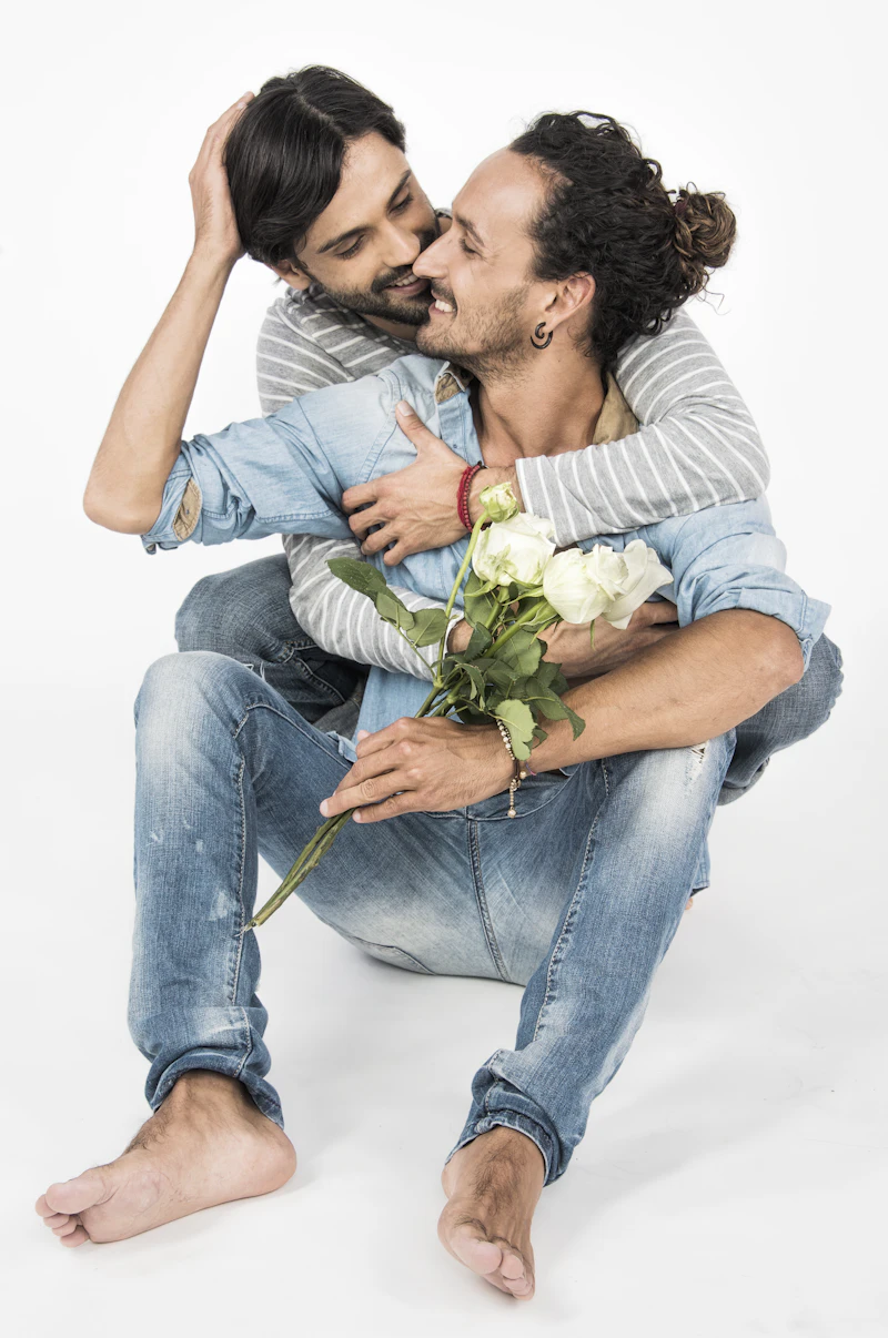 Two men embracing, one holding a small bouquet of white roses, both smiling
