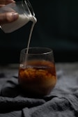 A hand is pouring milk from a small glass container into a larger glass filled with coffee. The milk swirls into the coffee, creating a mixing effect. The scene is set against a dark background, with the focus on the flowing milk and the cup.