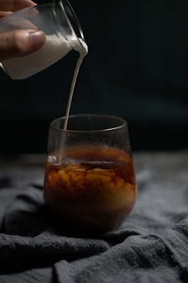 A hand is pouring milk from a small glass container into a larger glass filled with coffee. The milk swirls into the coffee, creating a mixing effect. The scene is set against a dark background, with the focus on the flowing milk and the cup.