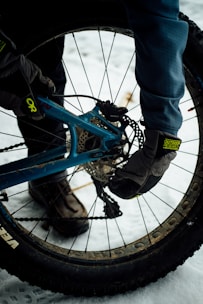 A person wearing gloves and winter boots is repairing or adjusting the rear wheel of a bicycle on a snowy surface. The bike has thick tires, suited for winter or off-road biking. The snow on the ground suggests a cold environment.
