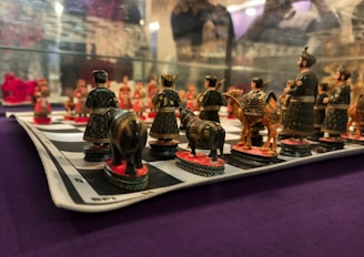 A detailed view of a chessboard with intricately designed pieces depicting figures in traditional attire. The pieces resemble soldiers, horses, and elephants, all meticulously crafted in dark and earthy colors. The chessboard is placed on a surface with a purple fabric background.