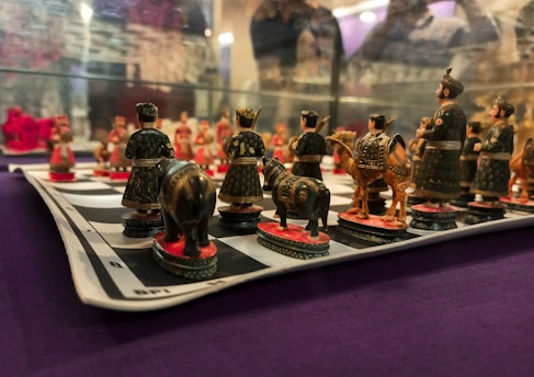 A detailed view of a chessboard with intricately designed pieces depicting figures in traditional attire. The pieces resemble soldiers, horses, and elephants, all meticulously crafted in dark and earthy colors. The chessboard is placed on a surface with a purple fabric background.