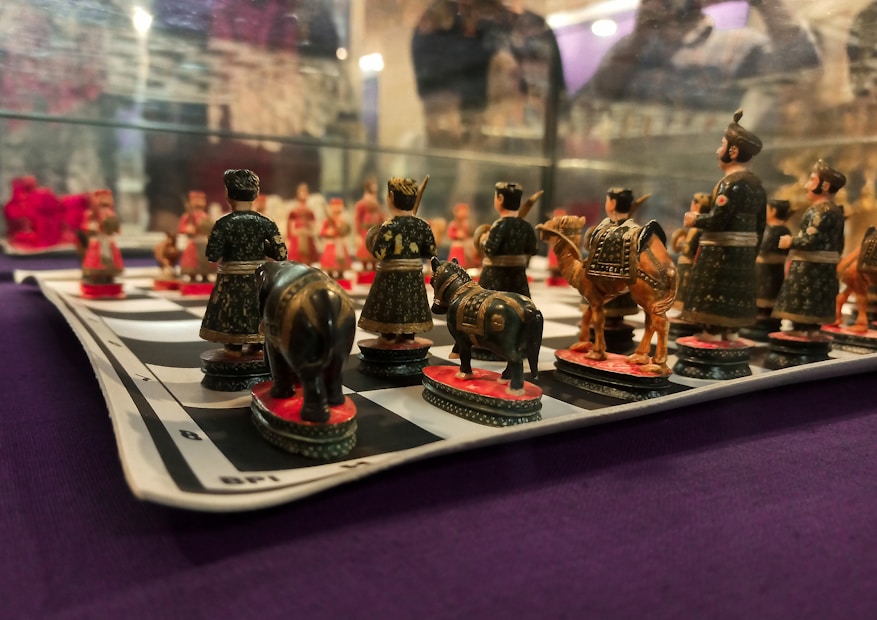 A detailed view of a chessboard with intricately designed pieces depicting figures in traditional attire. The pieces resemble soldiers, horses, and elephants, all meticulously crafted in dark and earthy colors. The chessboard is placed on a surface with a purple fabric background.