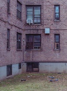 A brick apartment building with several windows, some having bars and others an air conditioning unit. The lower part of the building features basement windows with protective grates. In the courtyard area, a few pigeons are pecking at the sparse grass and fallen leaves.