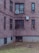 A brick apartment building with several windows, some having bars and others an air conditioning unit. The lower part of the building features basement windows with protective grates. In the courtyard area, a few pigeons are pecking at the sparse grass and fallen leaves.