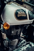 Close-up of a Royal Enfield motorcycle, focusing on the branding and parts of the engine. The logo is prominent on the bike's fuel tank, which is painted in a light color. Parts of the engine and other mechanical components are visible, with metallic textures and detailed craftsmanship.