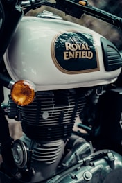 Close-up of a Royal Enfield motorcycle, focusing on the branding and parts of the engine. The logo is prominent on the bike's fuel tank, which is painted in a light color. Parts of the engine and other mechanical components are visible, with metallic textures and detailed craftsmanship.