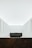 A minimalist interior space with stark white walls and ceiling. The room features a central dark-colored rectangular structure and another similar structure in the foreground. Subtle architectural details and lighting create a serene and spacious atmosphere.