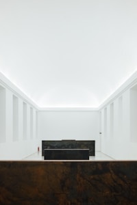 A minimalist interior space with stark white walls and ceiling. The room features a central dark-colored rectangular structure and another similar structure in the foreground. Subtle architectural details and lighting create a serene and spacious atmosphere.
