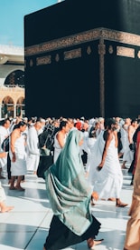 A large group of people walking around a black cuboid structure adorned with intricate gold patterns on its sides. The individuals are dressed in white robes, and a woman in a hijab is prominently walking in the foreground. The scene appears to be part of a significant cultural or religious event, taking place in a sunny, spacious plaza.